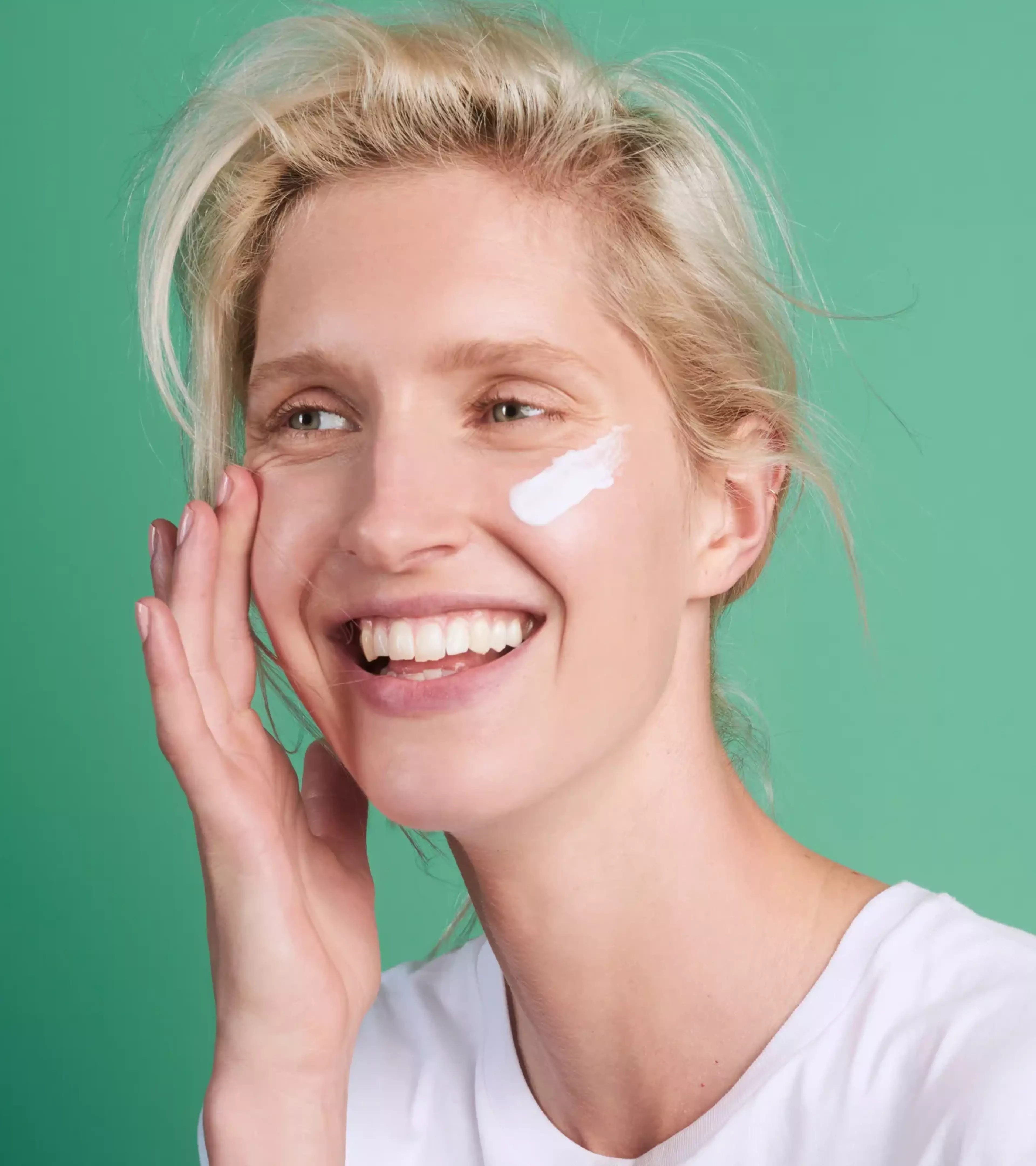 An image of a blonde woman applying cream to her face on a green background